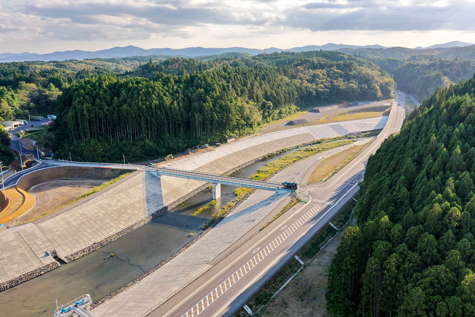 中央に橋がかかる川、両側に護岸と道路、右手には山が迫り、奥に向かって山間の道が続く、橋と護岸整備され山へと続く道路の写真