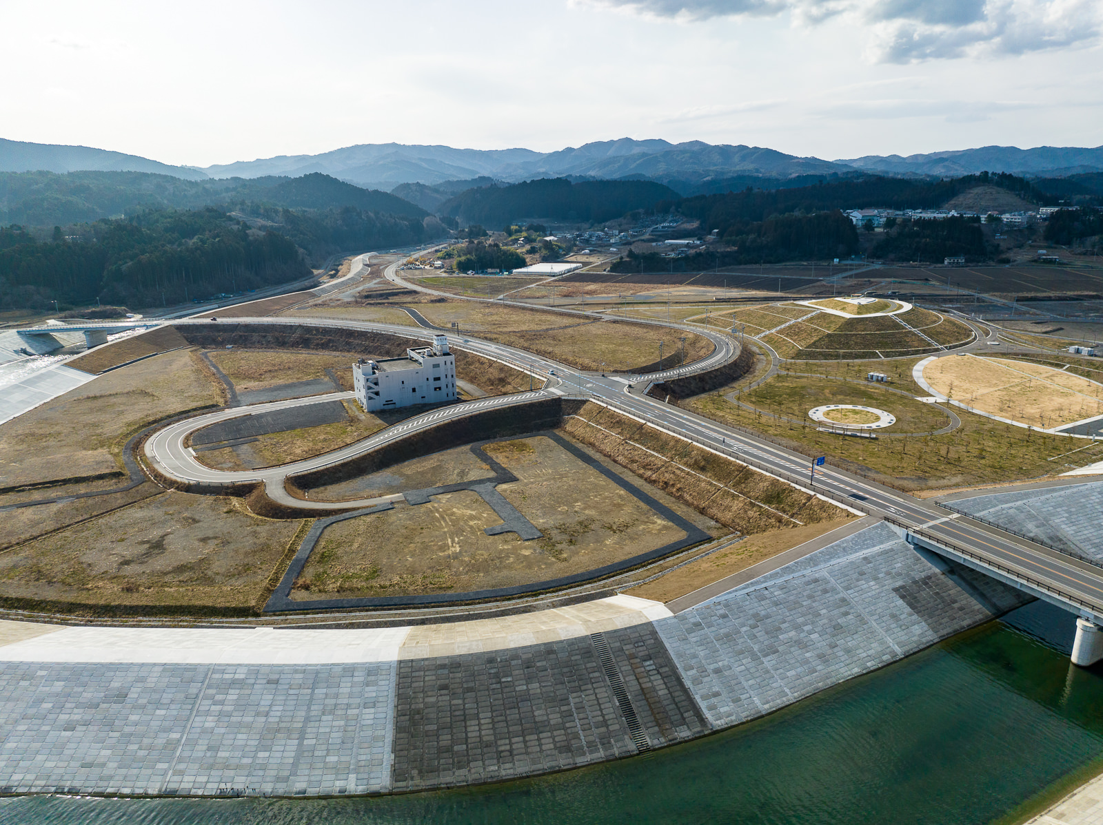 川を貫く道路の右側に緑地と円形の植栽がいくつかあり、左側には白い建物があり、背後には山並みが続く、全体が整然と整備された復興公園付近の昼間の風景を高台から定点カメラで撮影した写真