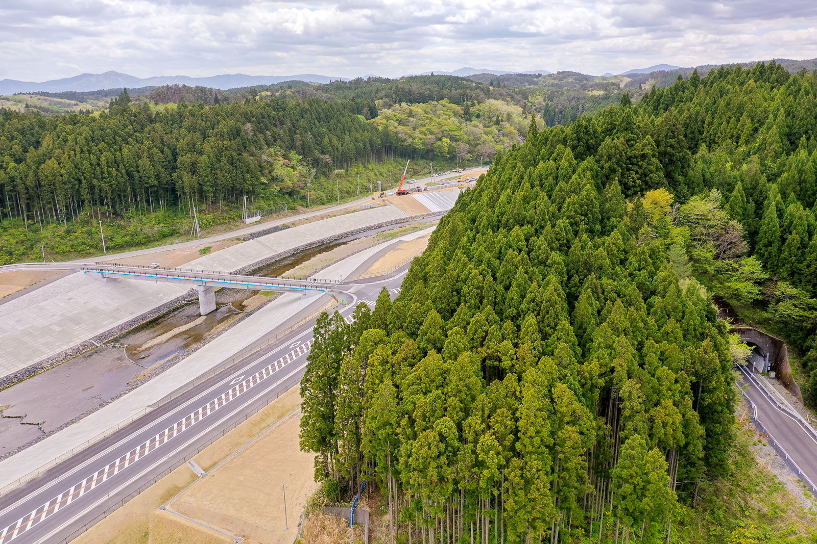右側に広がる密集した森林と、左側に建設中の高架道路が乾いた川のような地形をまたぎ、背景に工事車両と緑の丘陵が広がる風景の写真