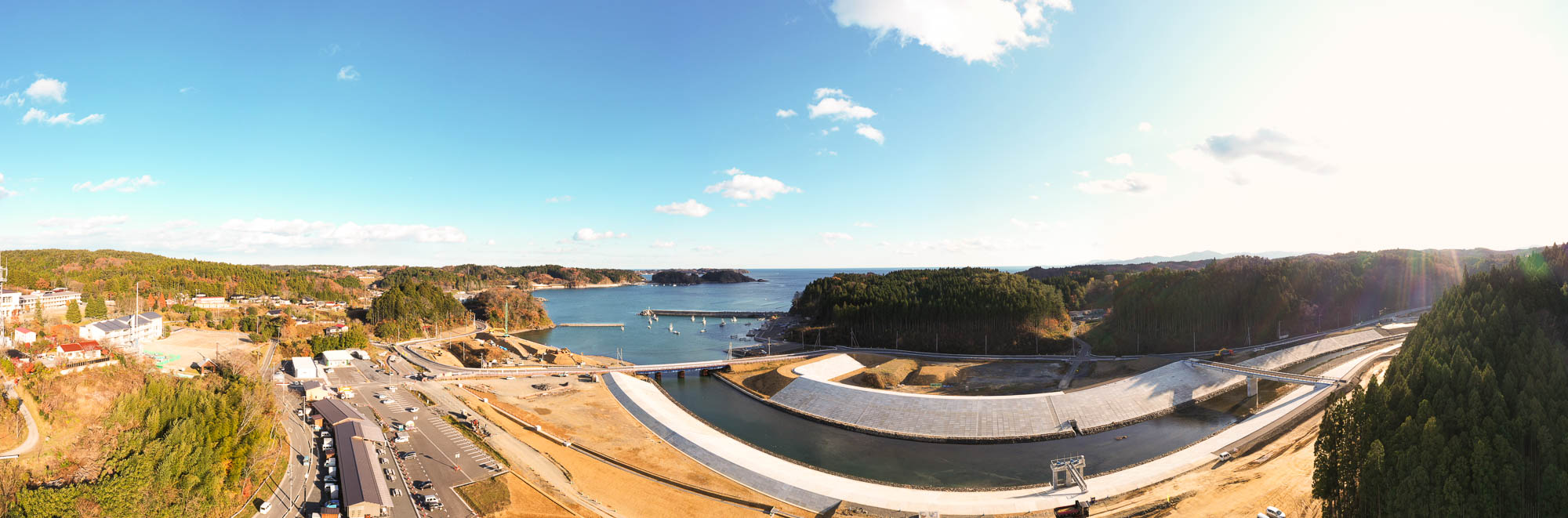 湾に面した港町の全景を高所から広く見渡しており、左側に町並みと駐車場、中央に港と防波堤、右側には川と整備中の護岸が森の中を蛇行して続いている様子を写した写真