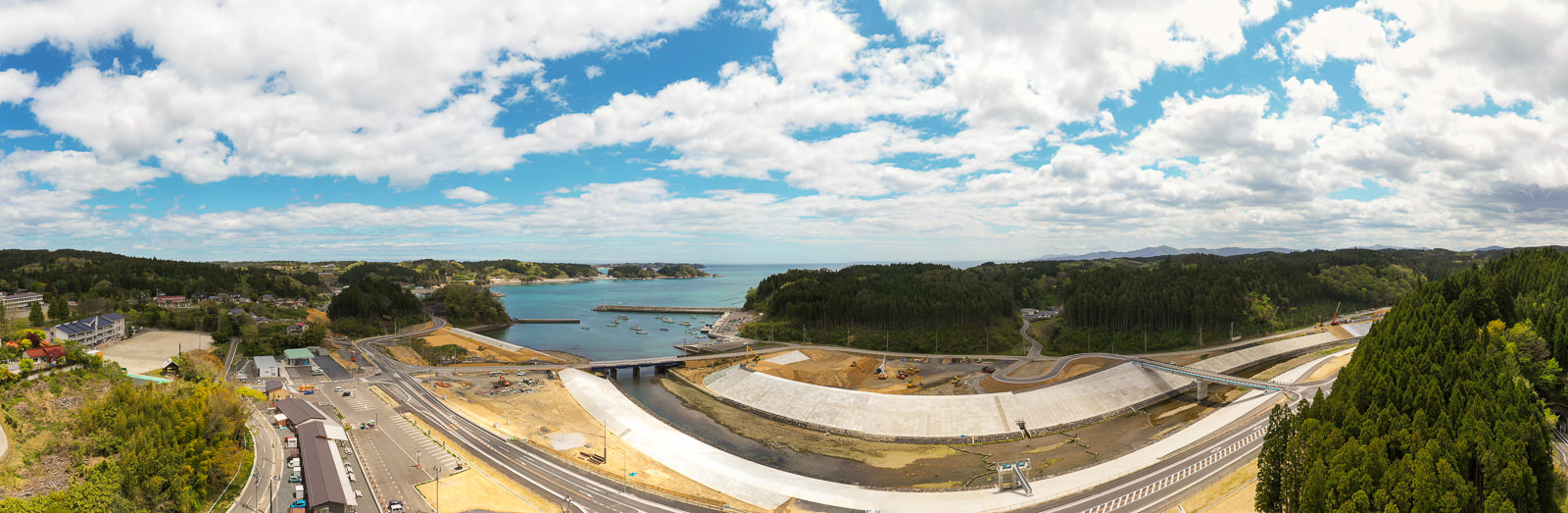 中央に入り江のような水域が広がり、周囲を緑の丘や森林が囲み、手前には道路や橋、大型のコンクリート構造物が並ぶ工事中の海辺の地域の写真