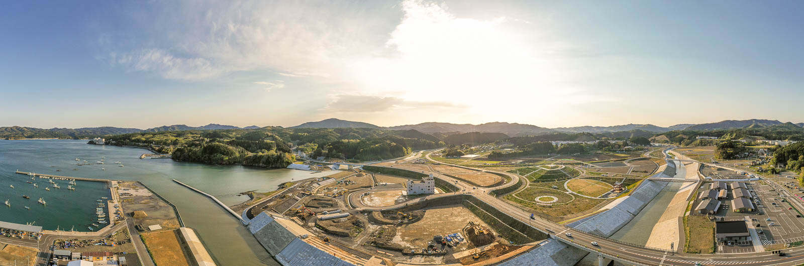 左に漁船が停泊する港と青い海、中央に川とその周辺で進行中の大規模な造成地、右には整備された道路と建物群、背景には広がる山々と空に太陽が輝く様子を、広角でパノラマ撮影した再開発中の港町全景の写真