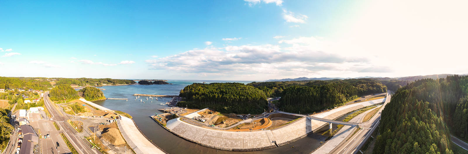 青空の下、右手に湾と防波堤、左手に山や集落、中央に川とそれに沿うように整備中の道路と橋が見える全体俯瞰の写真