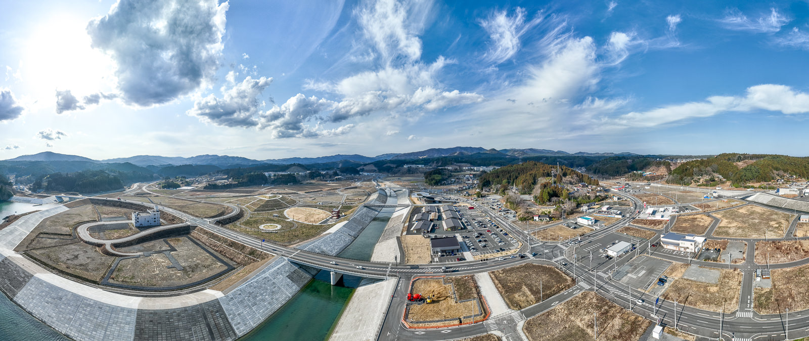 広範囲にわたる復興整備地域が写っており、中央には川と護岸がまっすぐ通っており、川の両側には道路と区画整理された空き地が広がり、奥には山々が連なる、復興工事が進められている町の全体の昼間の風景を高台から定点カメラで撮影した写真