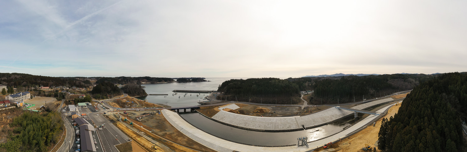 晴れた空の下、湾のような広い水域に面した海沿いの地域で、左側には道路網と複数の車両や建物が見え、中央から右側にかけてはダムや治水施設と思われる大規模なコンクリート構造物を含む工事現場が広がり、周囲は森林に覆われた丘に囲まれている沿岸開発地域の写真