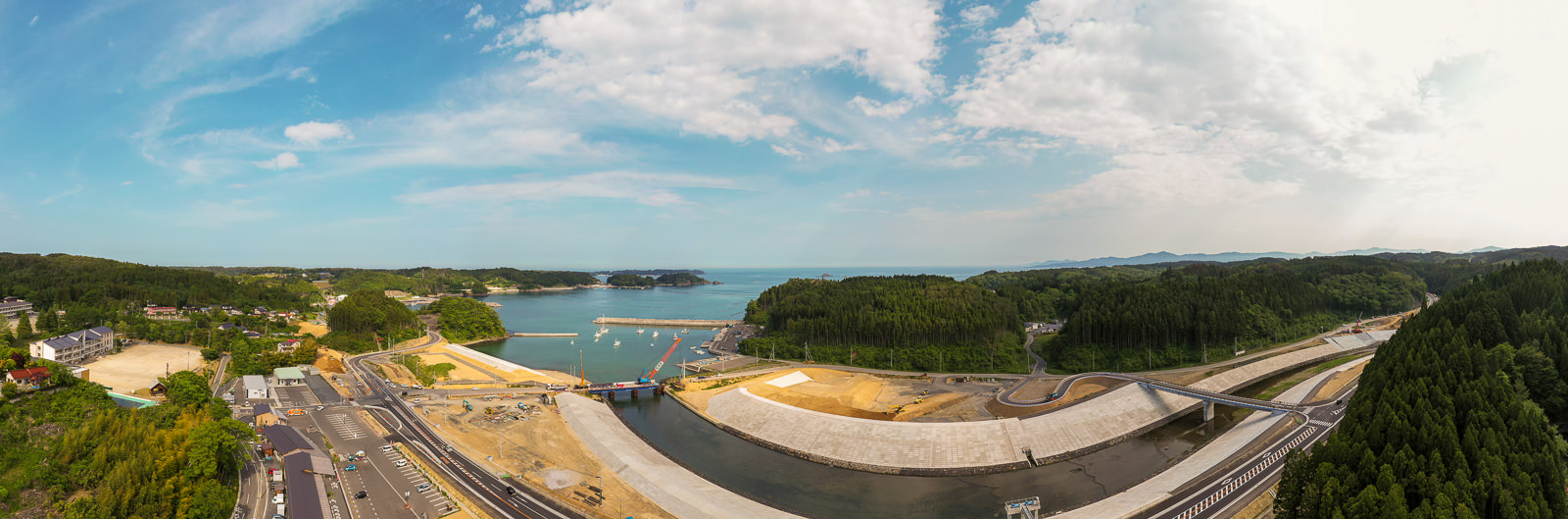 中央に水域が広がり、左側に建物と道路、右側に大規模なコンクリート構造物と道路の建設が進むインフラ整備中の沿岸地域を、緑の丘と森林に囲まれた晴れた空の下の写真