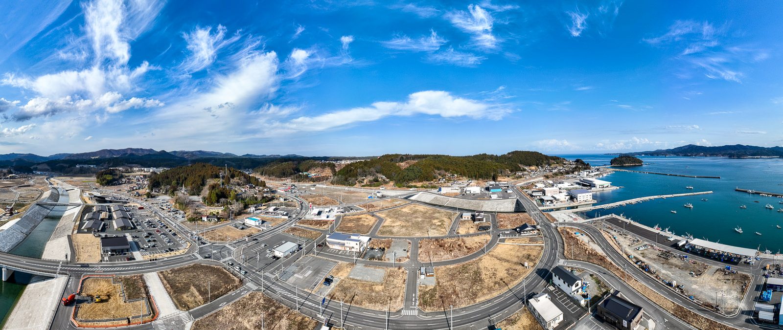 右側に広がる海と港、左側には整備された川と区画整備が進む土地が一体となって写されており、復興による都市の再構築と港町の風景が広がる港と街並み全体の昼間の風景を高台から定点カメラで撮影した写真