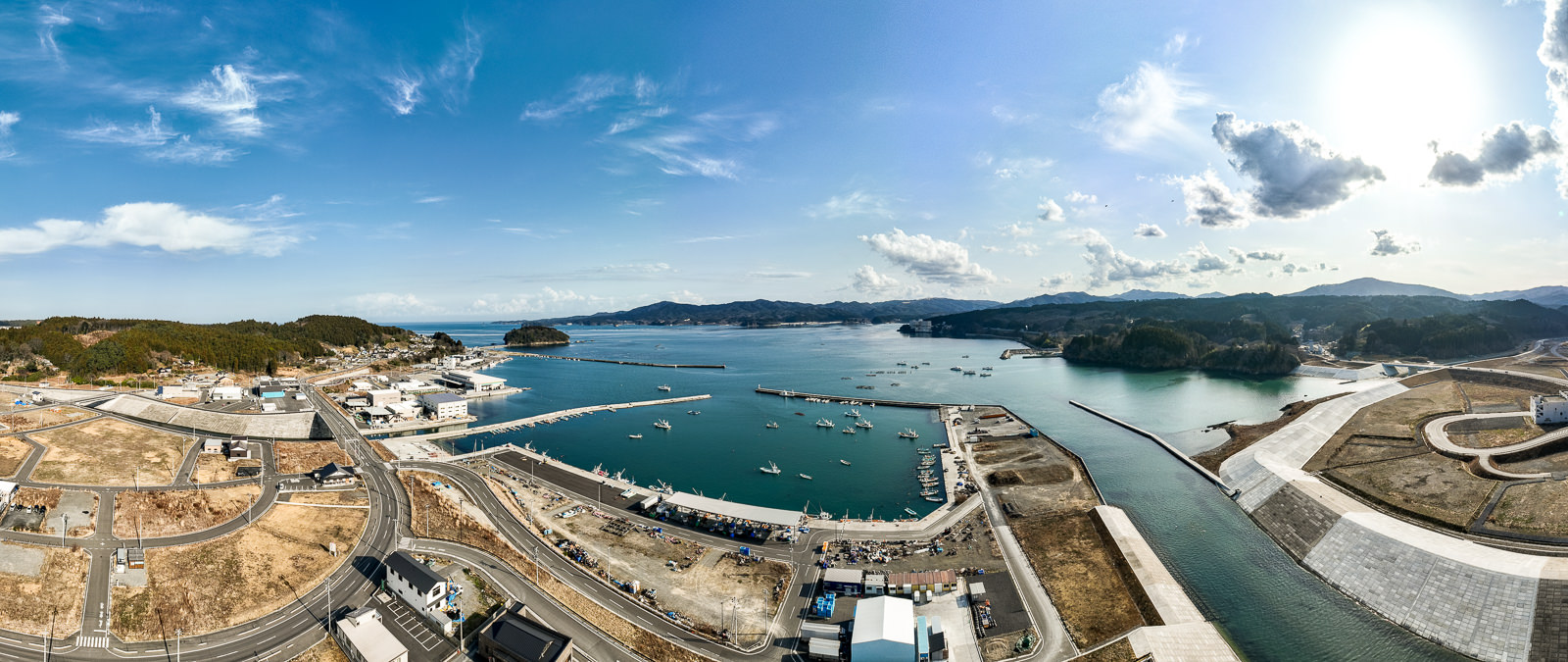 防波堤に囲まれた湾内には多くの船が浮かび、港周辺には倉庫や建物が建ち並ぶ、湾と漁港の昼間の風景を高台から定点カメラで撮影した写真