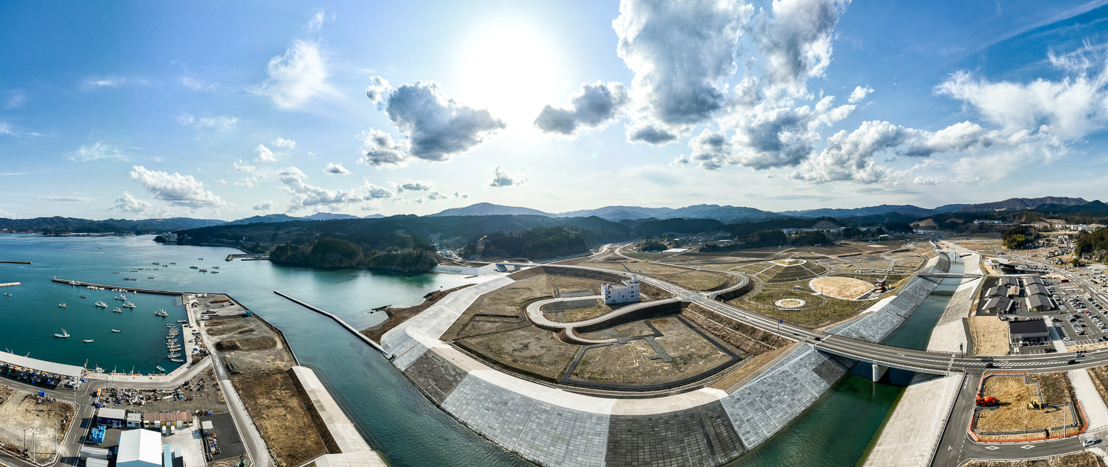 湾と川が交わる地点から見た全体風景で、左右に防波堤と護岸が延び、中央に川がまっすぐ流れ、両側には都市再開発された土地と緑地、道路が整然と配置されている復興都市全体の昼間の風景を高台から定点カメラで撮影した写真