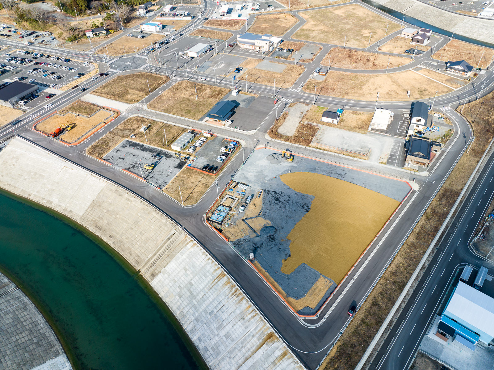 河川に面した広い護岸工事区域のすぐ隣に整備された道路と区画整理された土地があり、その中には工事中の建物や駐車された車両がいくつか見え、まだ建物の少ない地域が新しく整備されつつある復興途中の街並みの昼間の風景を高台から定点カメラで撮影した写真