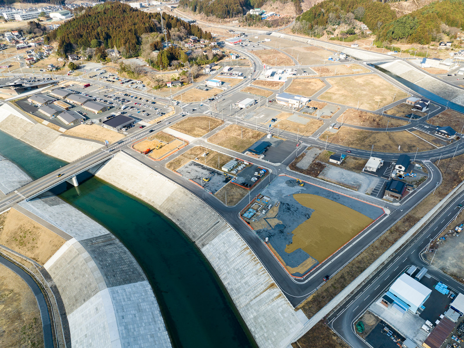 川に架かる橋を中心に広範囲にわたって道路や区画整理された空き地が広がり、その中に数軒の建物や車、建設作業中の場所が点在しており、周囲には山や林が見え、自然に囲まれた地域で復興や開発が進んでいる昼間の風景を高台から定点カメラで撮影した写真