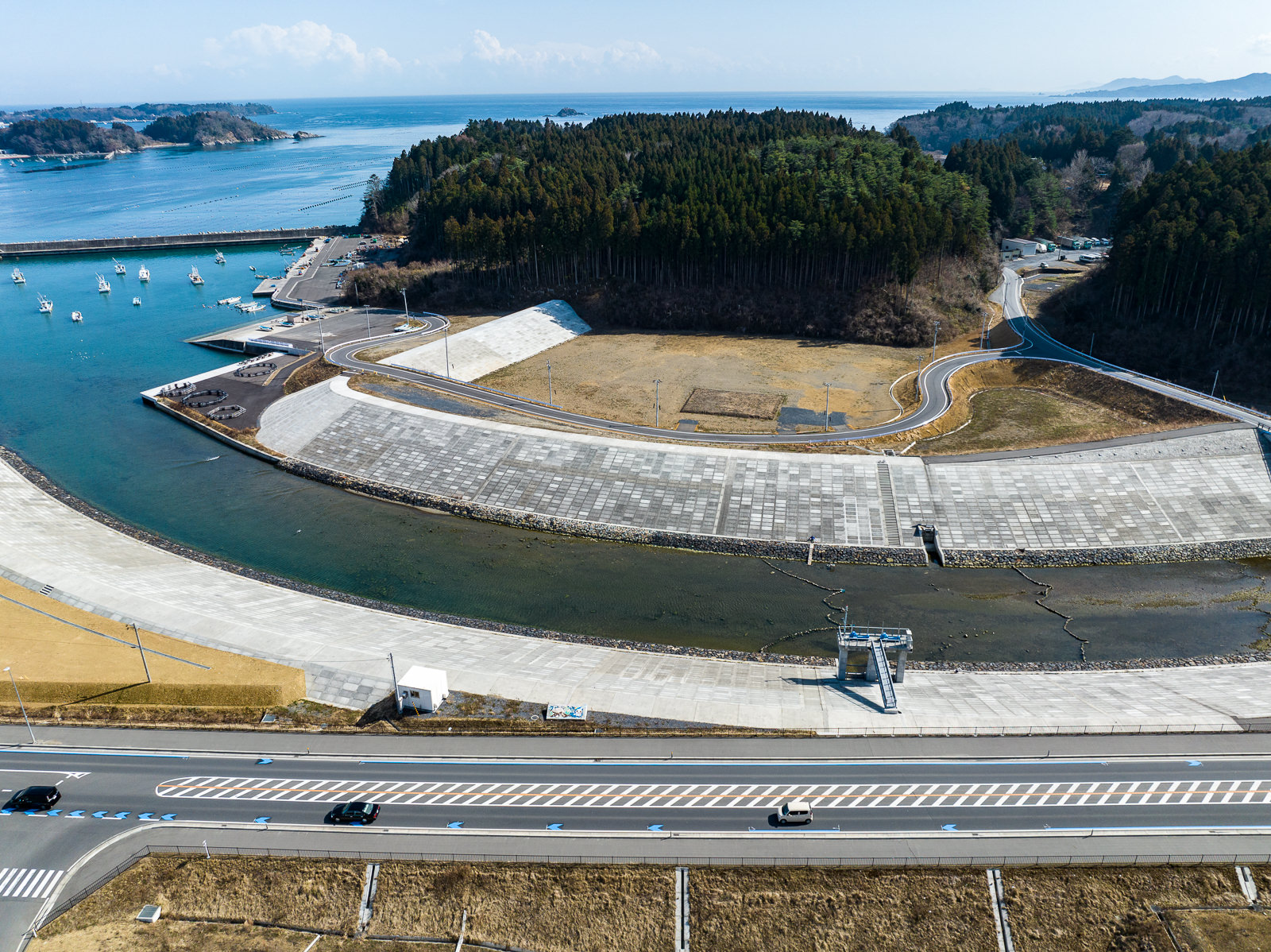 護岸工事が施された川が海へと流れ込み、湾内に停泊する漁船とそれを囲む堤防、道路が湾曲しながら海へ続く様子を示す港湾周辺の昼間の風景を高台から定点カメラで撮影した写真