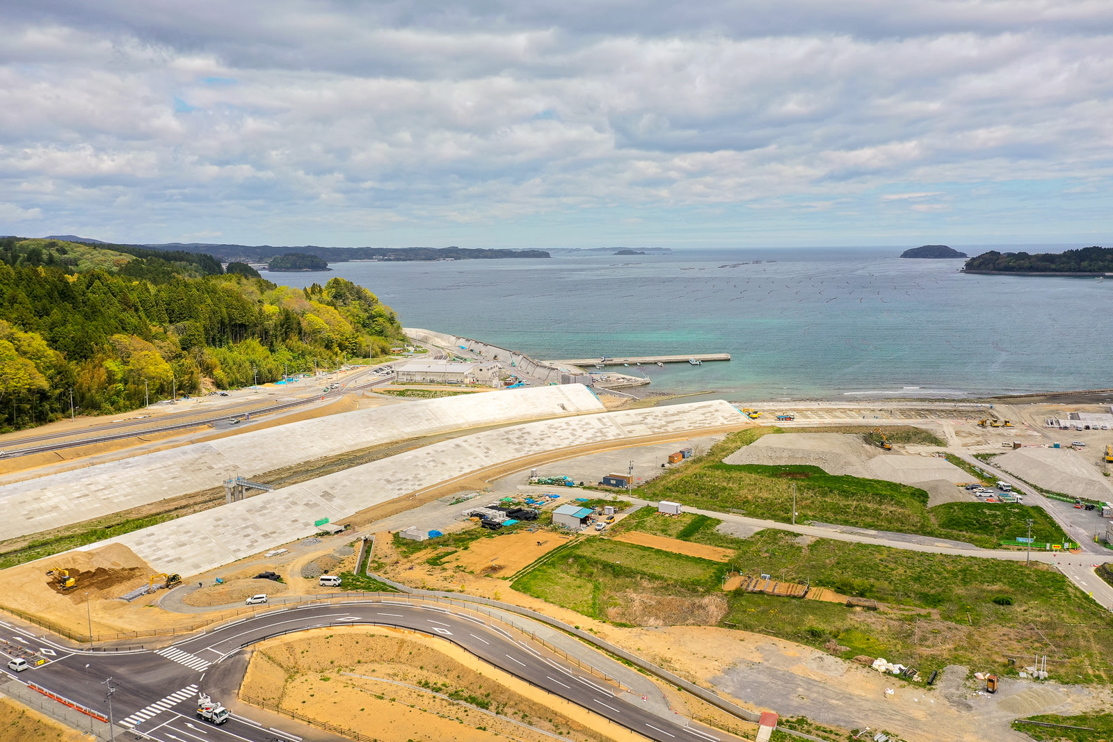 海岸沿いに大きな防波堤と緑の草地、曲がりくねった道路が広がり、遠くの海には小さな島々が見え、周囲には建設車両や機材が点在する沿岸地域の写真