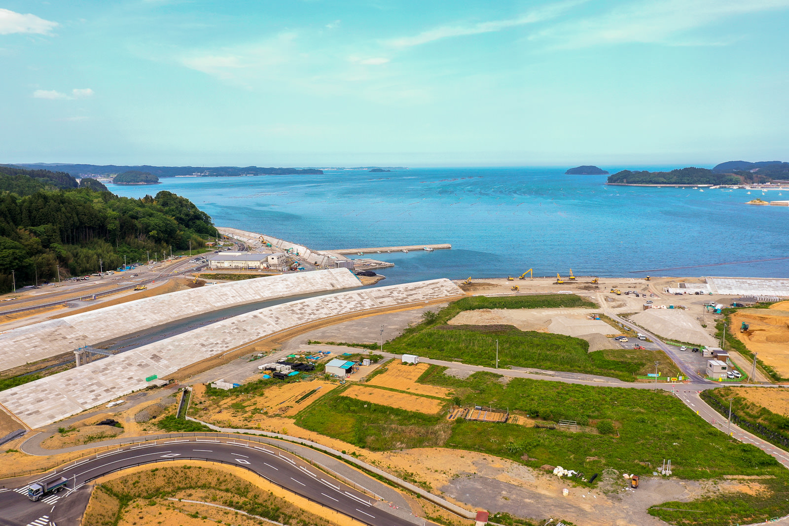 海岸線に沿って大きな防波堤が築かれ、草地や開発中の土地が広がる区域を曲がりくねった道路が通り、背景には青い空と遠くの陸地が見える海沿いの地域の写真