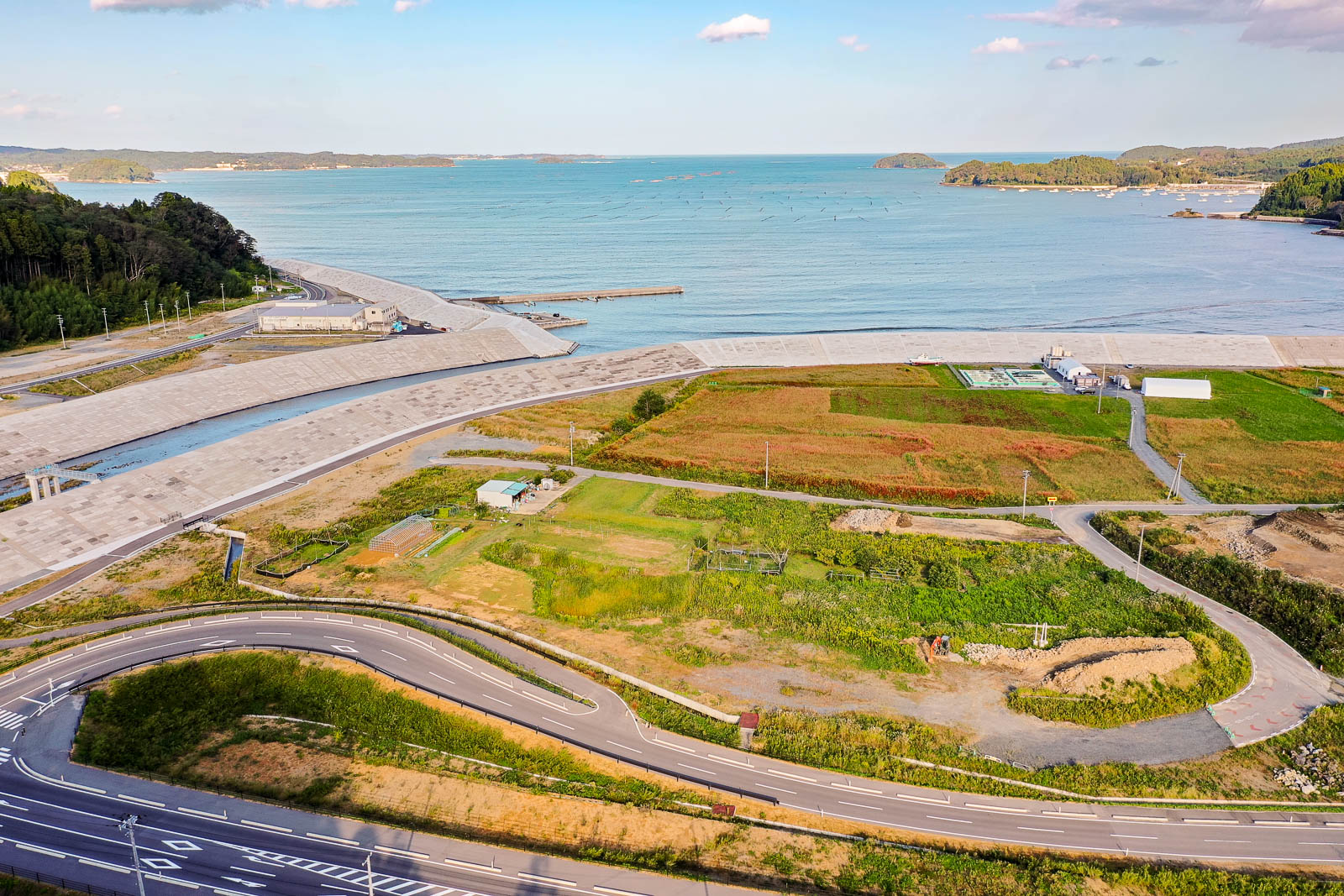 防潮堤と海のあいだにある農地や小さな建物が点在する地域を、海に向かって見下ろす構図で撮影されており、沖合には養殖らしき設備が並ぶ様子も確認できる写真