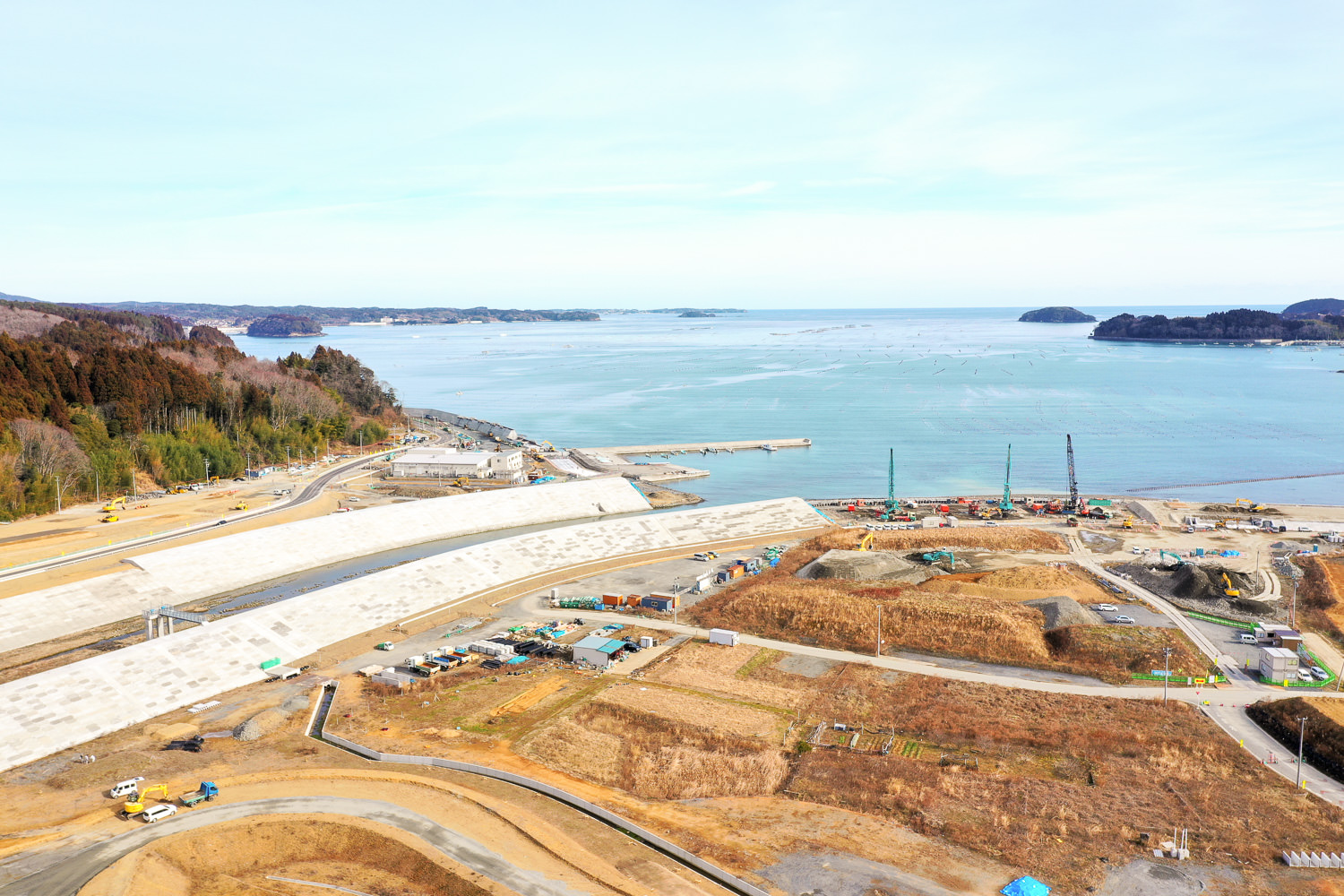 海岸沿いの工事現場で、大型のコンクリート壁や道路、複数のクレーンが配置されており、背景には海と小さな島々や陸地が見え、造成された土地には仮設の建物やコンテナが点在している写真