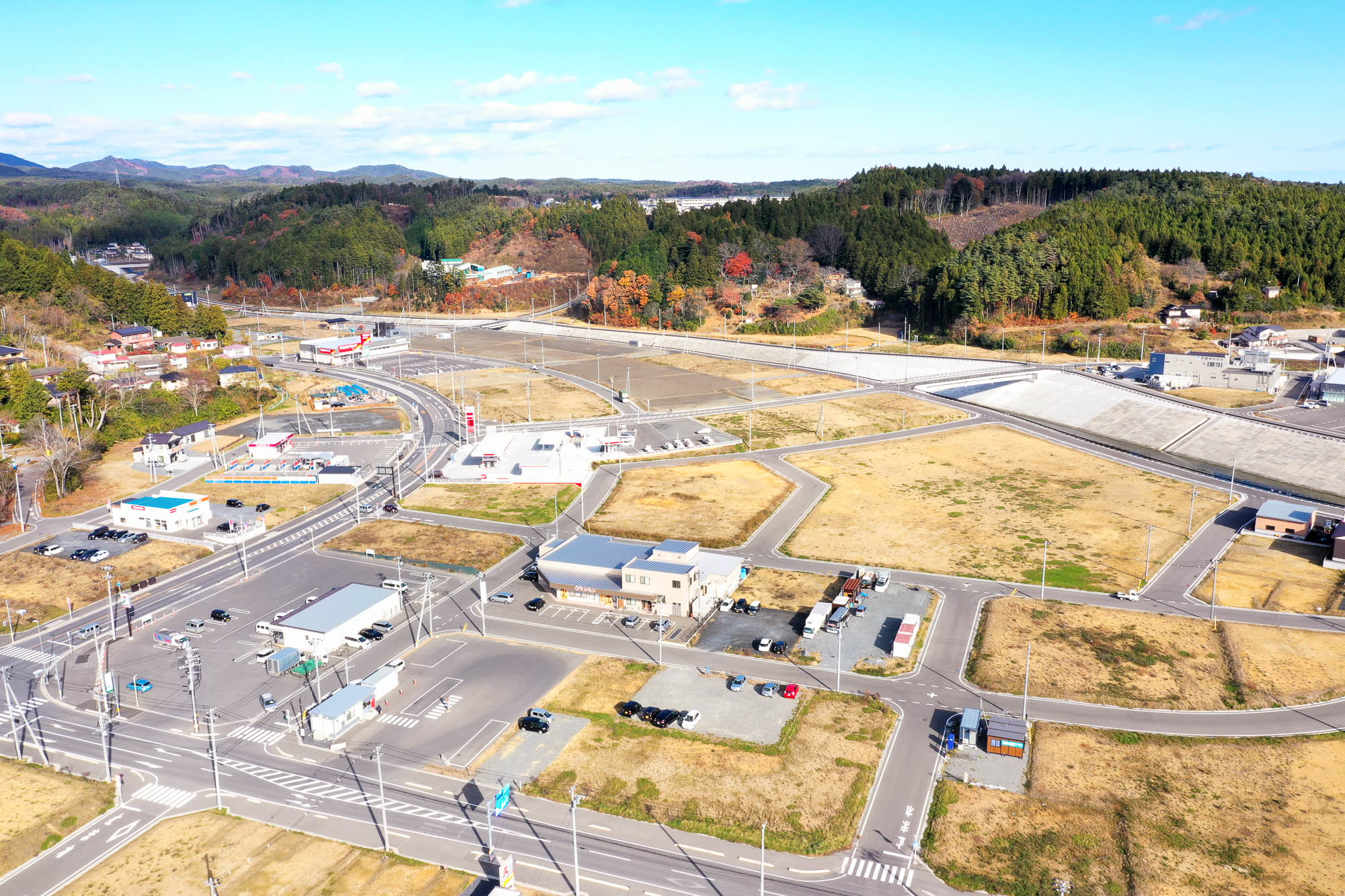手前の交差点から奥へと続く造成地の道路と空き地の配置、周囲の商業施設や山々を見下ろした様子の写真