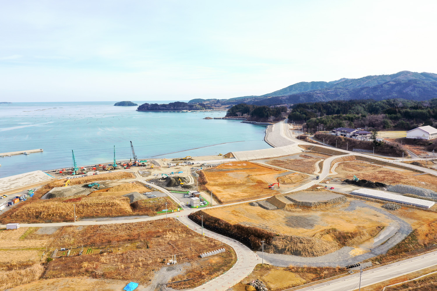 晴天のもと、湾に沿って湾曲した海岸沿いの地域で、複数のクレーンや建設機材が稼働する工事現場が広がり、道路や通路が点在し、背景には丘や森林が見える写真