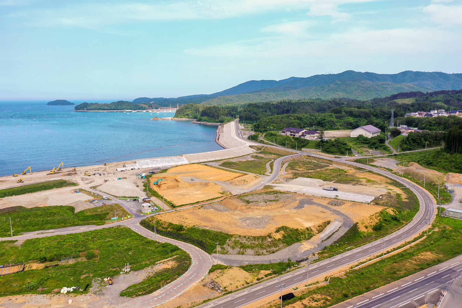 左側に水辺が広がり、緑の草地といくつかの工事現場を曲がりくねった道路が通り抜け、背景には丘や山々が連なり、空には雲が浮かぶ自然と開発が共存する海沿いの地域の写真