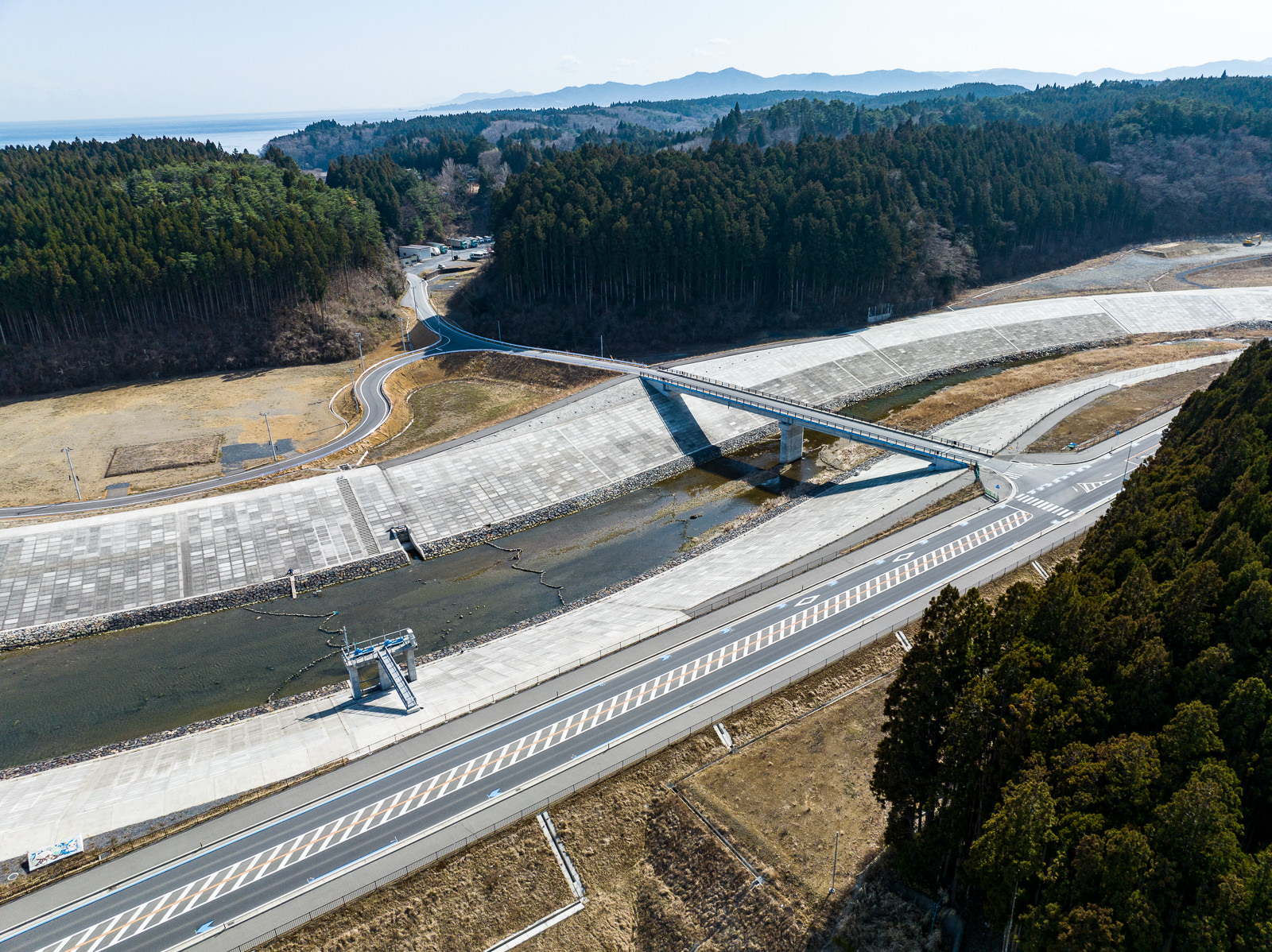 大きな護岸コンクリートで囲まれた川にかかる橋と、川沿いの道路や山へと続く道が見え、全体に護岸工事が施されている昼間の風景を高台から定点カメラで撮影した写真