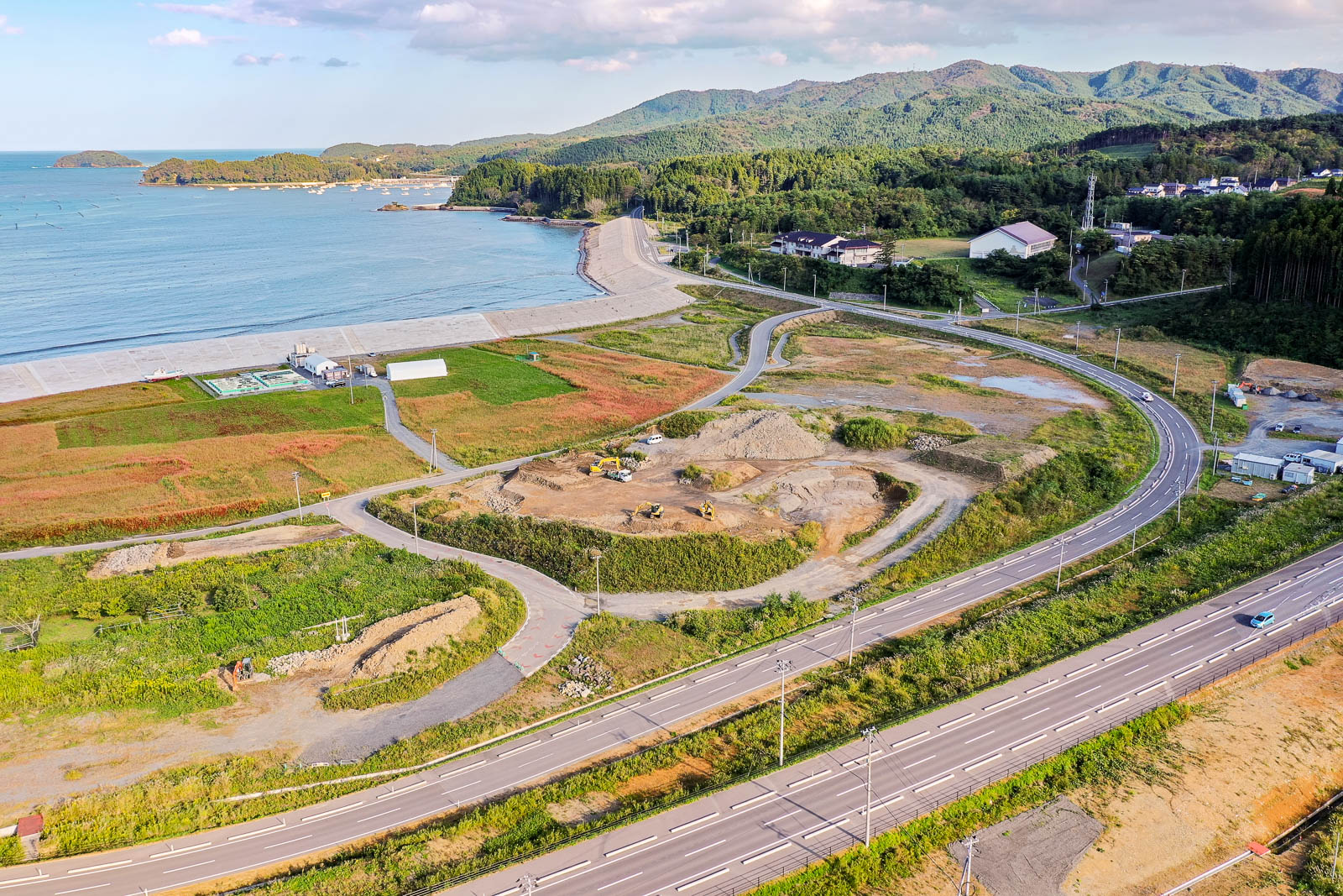 海と接する湾曲した堤防に沿って整地された広い土地と建設中の重機が並ぶ区画、その背後に小高い山と住宅や学校らしき建物が見える、再開発が進む沿岸地域の写真