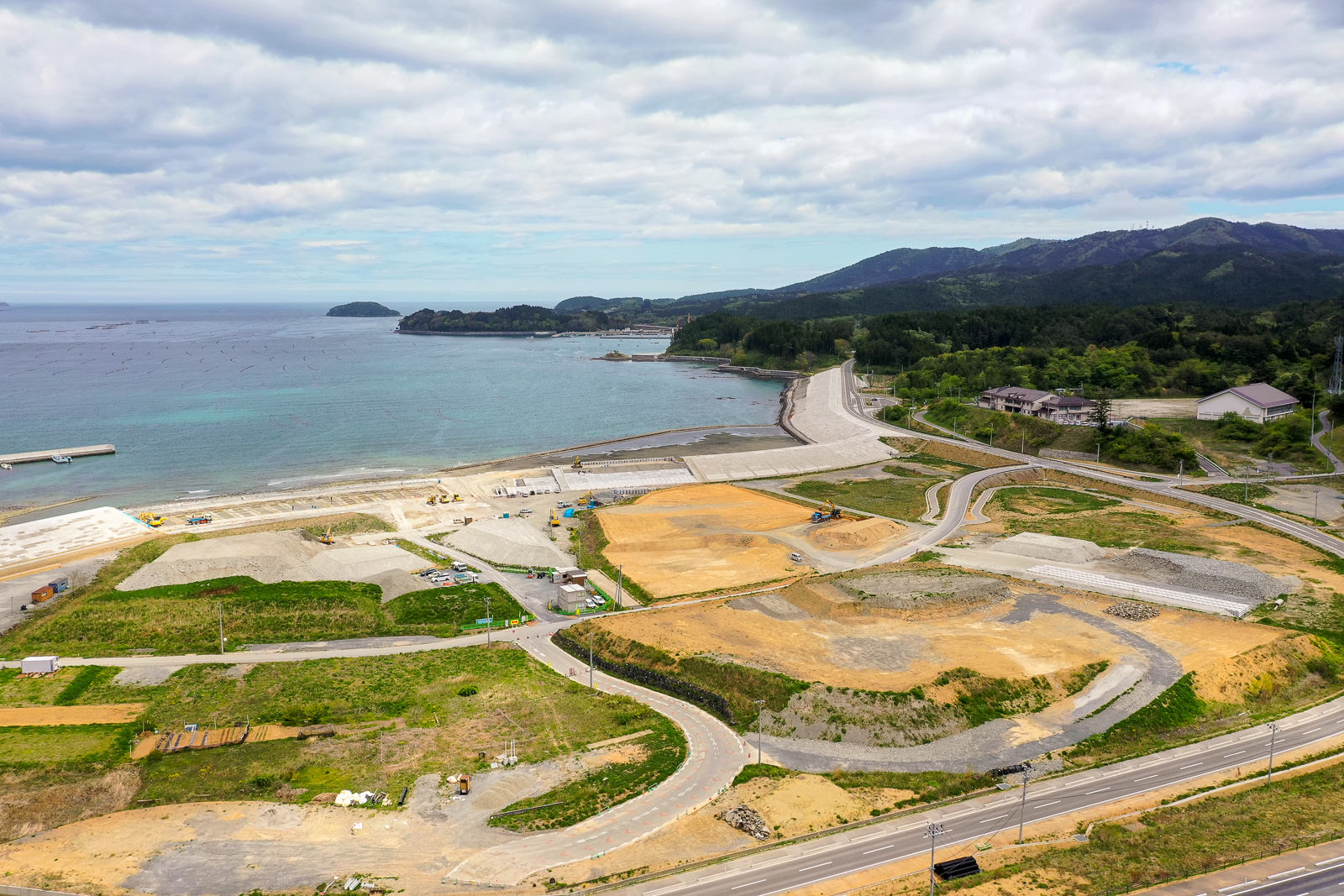 海岸沿いに整備中の土地と道路が広がり、その奥には緑に覆われた山々と穏やかな海が見える、自然と開発が共存する風景を写した沿岸地域の写真