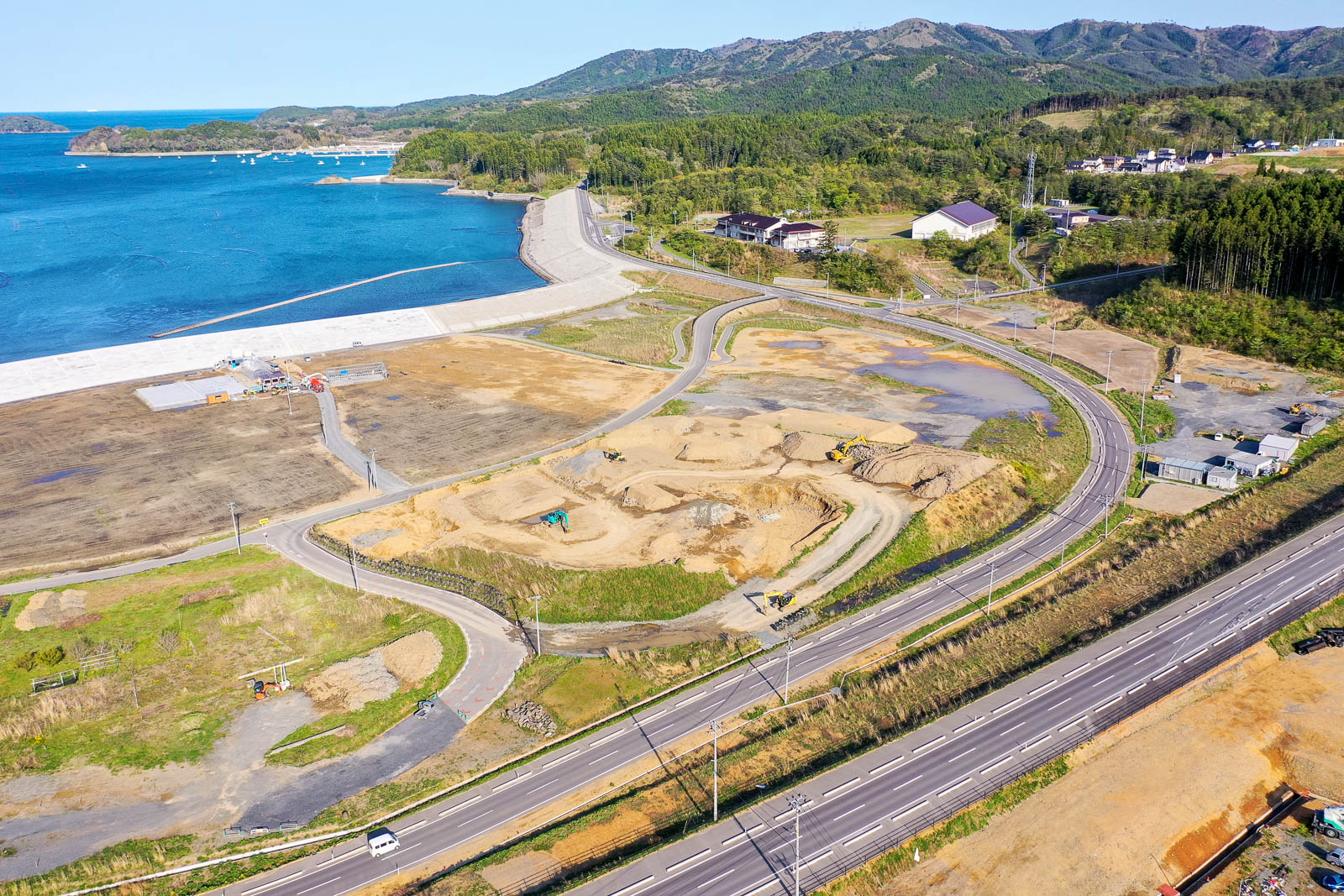 手前には複数車線の整備された道路が走り、その奥に造成中の土地が広がって重機が稼働しており、盛土や整地作業が進められている写真