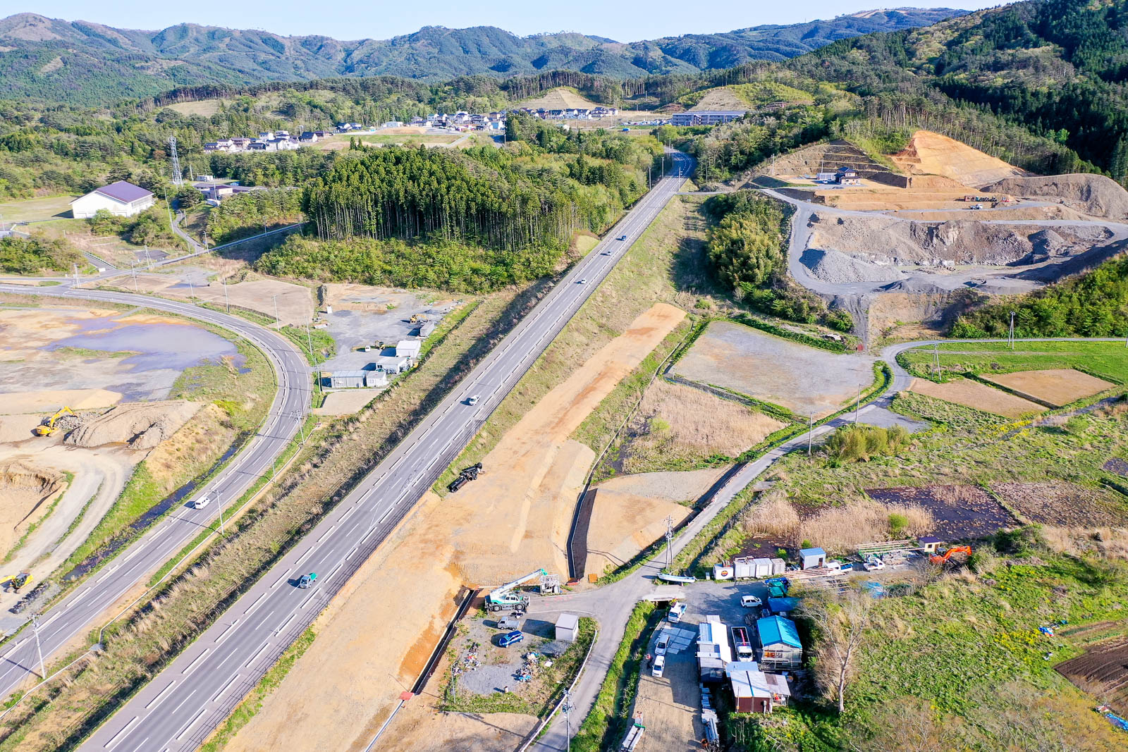 中央には片側2車線の主要道路が真っすぐ延び、その両脇では斜面の切土や盛土工事が行われ、右側の山肌は大きく削られ、砕石や土砂が積み上がっており、造成作業が進行中であることがうかがえる写真