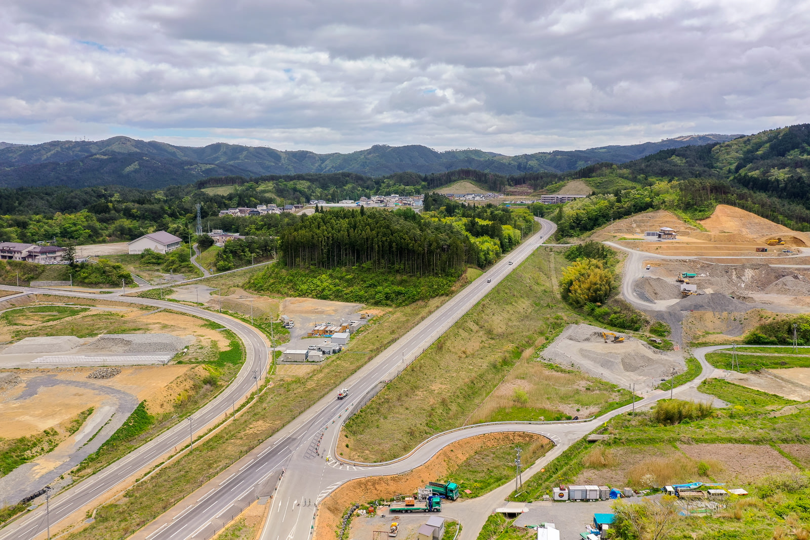緑に囲まれた土地に複数の道路が張り巡らされ、背景には丘が広がり、所々に工事現場と建設車両が見られ、空には雲が浮かぶ開発中の地域の写真