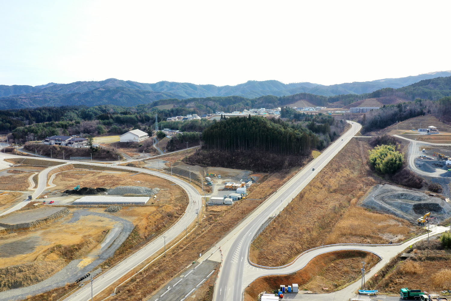 緑豊かな山々と森林を背景に、広大な土地が広がる風景を上空から捉えたもので、中央には舗装された道路が右方向へと伸びており、その両側には未開発の土地や建設中のエリアが広がっている。左側には曲がりくねった道が見え、いくつかの建物や構造物が点在しており、全体として開発が進行中の地域の様子がうかがえる写真