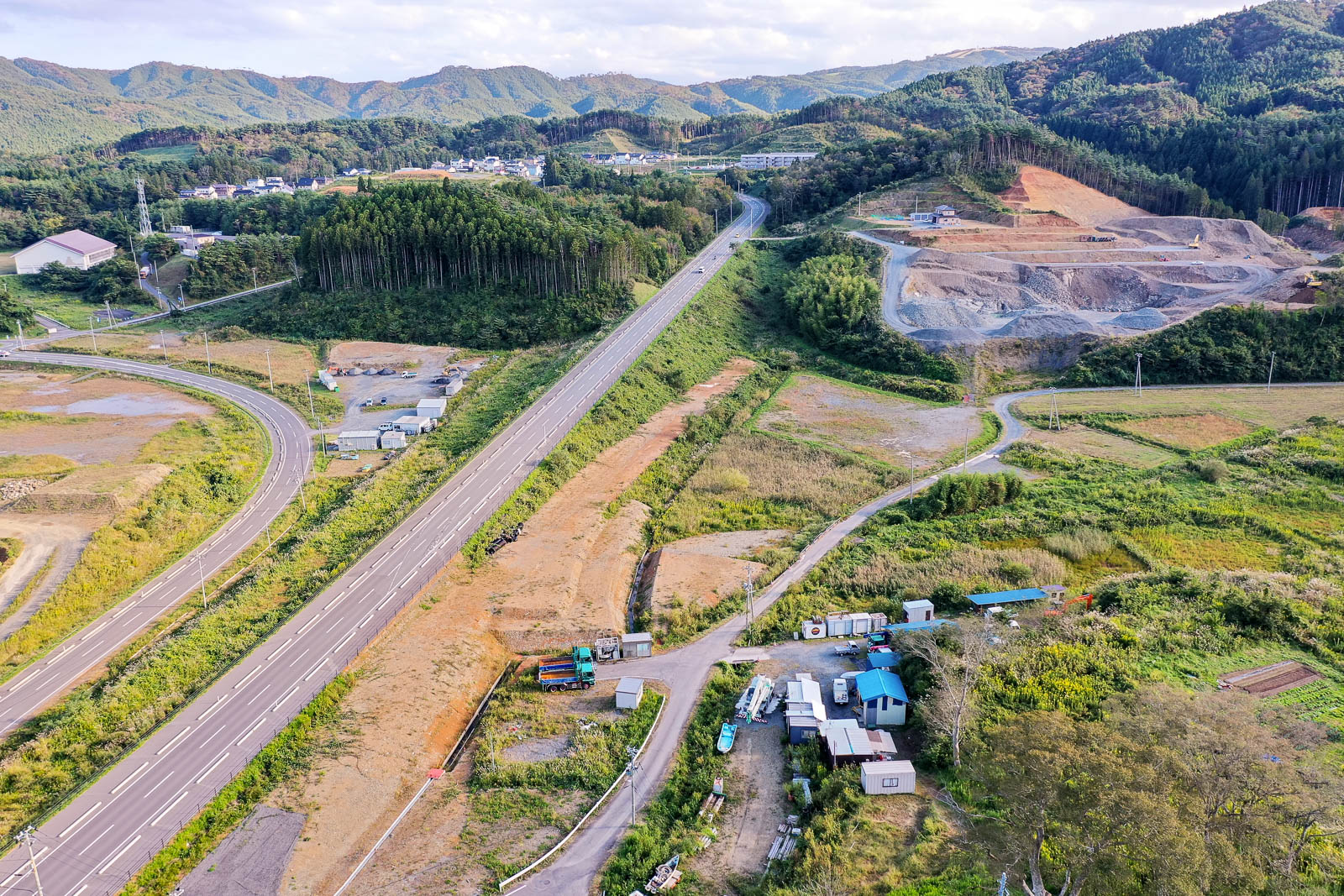 山間の道路が一直線に延び、その両側に木々が生い茂るエリアと造成中の土地が広がっていて、手前には建設機材や小さな建物が密集している様子が確認できる写真