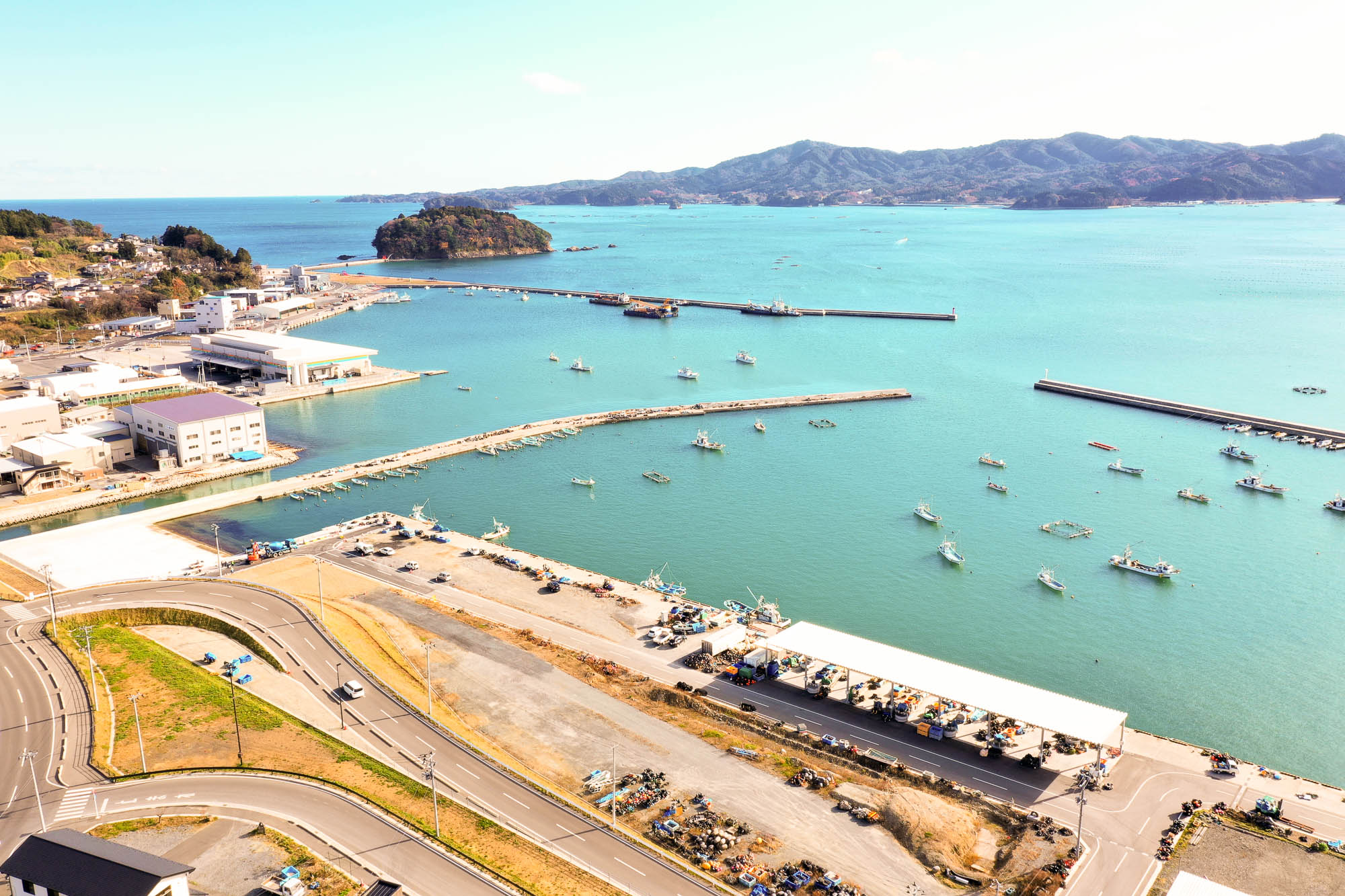 穏やかな青い海に多くの漁船が浮かび、手前には道路と倉庫、左側には工場や建物が並ぶ様子が見える漁港の左側から見た写真