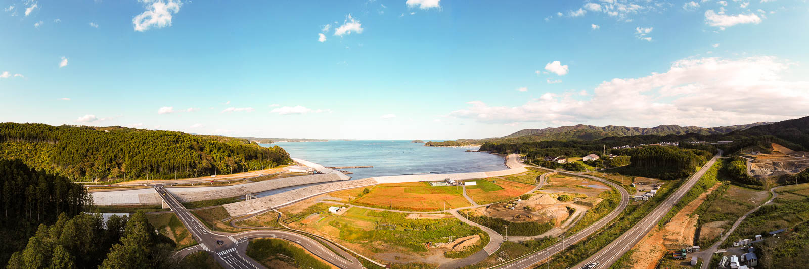 海に面した湾の内側を大きく囲うように整備された堤防と、その内側に広がる整地された土地や道路、新しいインフラが整備されつつある様子を遠くの山並みと一緒に捉えた写真