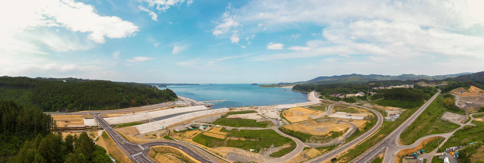 複数の広い道路が緑地や建設中の区域を通り抜け、背景には山々と青空が広がる自然と開発が共存する海岸沿いの地域の写真