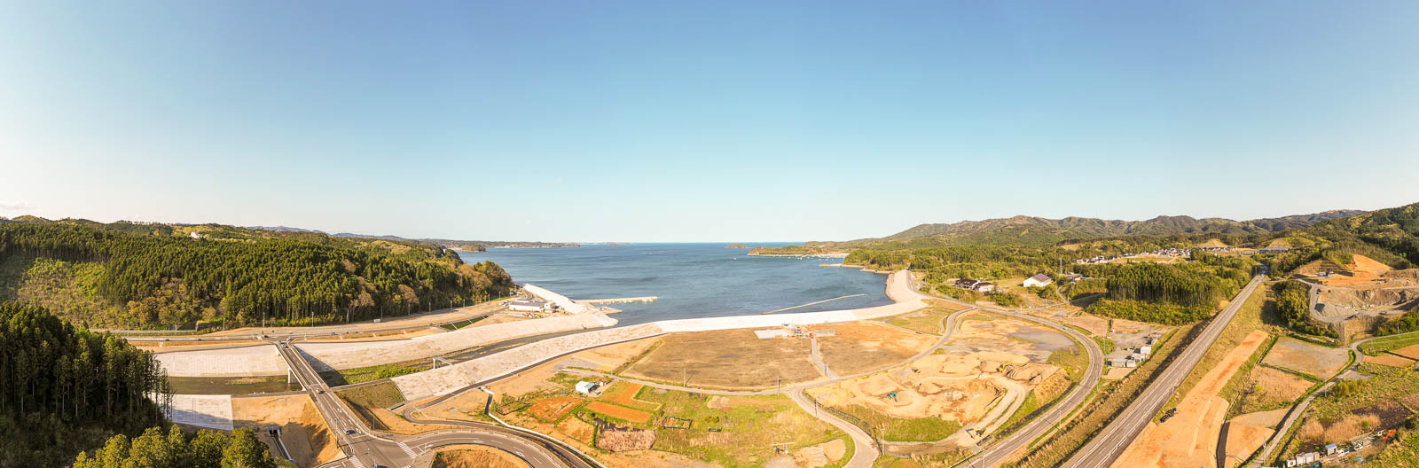 中央奥には広がる海、その手前には真新しいコンクリート製の防潮堤や護岸が長く続いています。左奥と右奥の両サイドから堤防が海を囲むように伸び、港の入り口を形成してるパノラマ写真