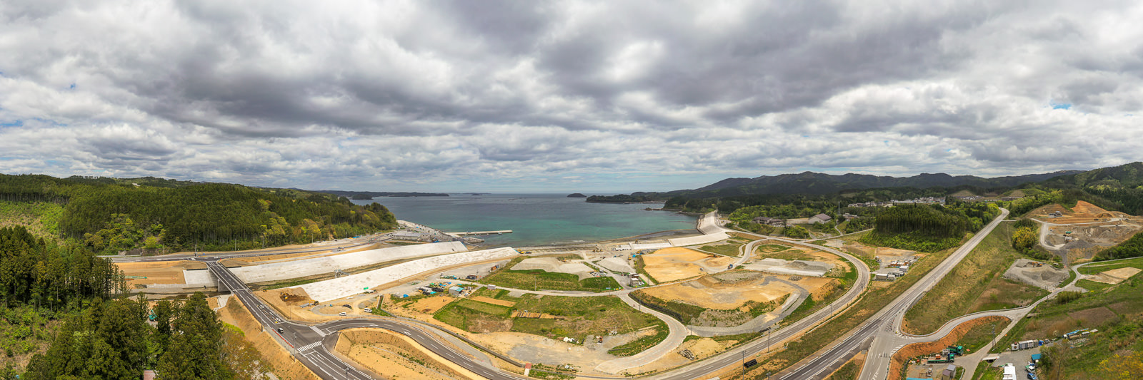海に面した地域を広く見渡すパノラマ写真で、道路や工事現場、緑地が点在し、海岸線の向こうには丘と森林が広がり、空には雲がかかっている沿岸地域の写真