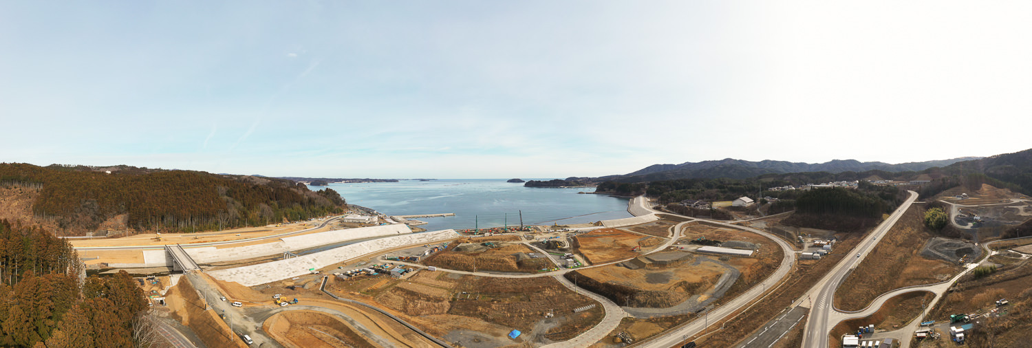 広い湾に面した海岸沿いの地域で、複数の道路が交差しながら建設中の構造物や工事現場が点在している様子を広く捉えた写真