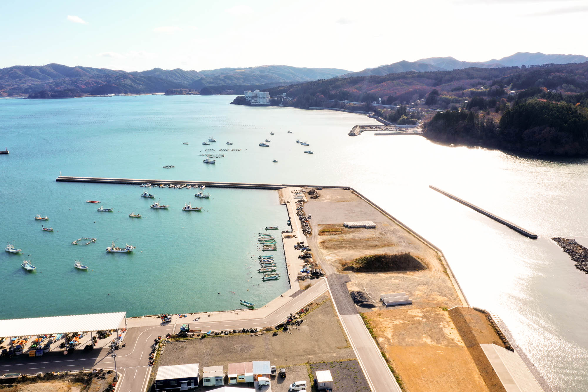 防波堤に囲まれた湾内に漁船が集まり、右奥にはリゾートホテルのような白い建物が見え、湾の奥には山々が連なる漁港の右側から見た写真