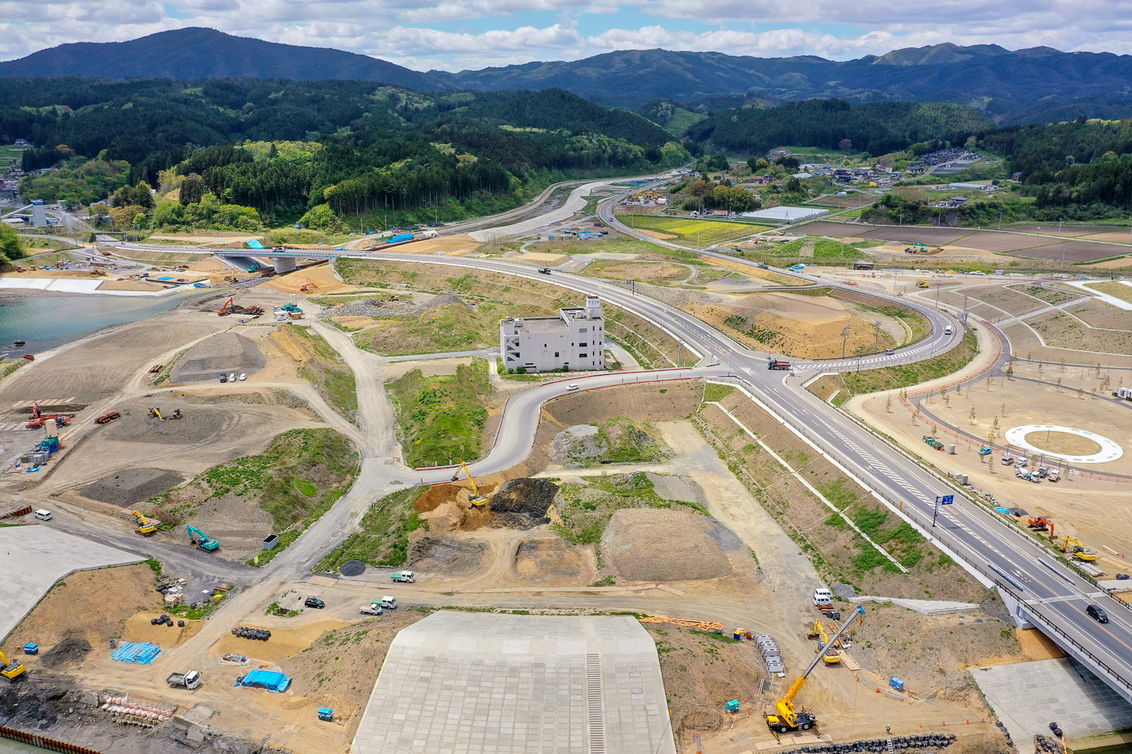 複数の道路や通路が交差し、建物や工事車両が点在する広大な工事現場を上空から撮影し、背景に緑の丘陵が広がる写真