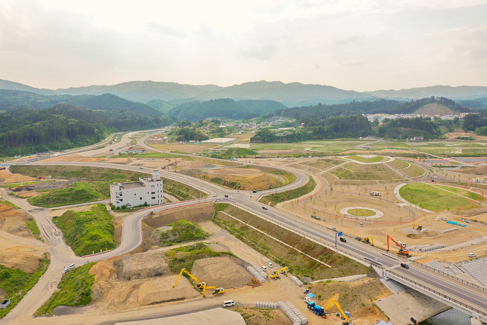 複数の重機や車両が稼働する大規模な建設現場で、道路や橋、円形の構造物が建設中であり、背景には緑に覆われた山々と点在する建物が見える写真