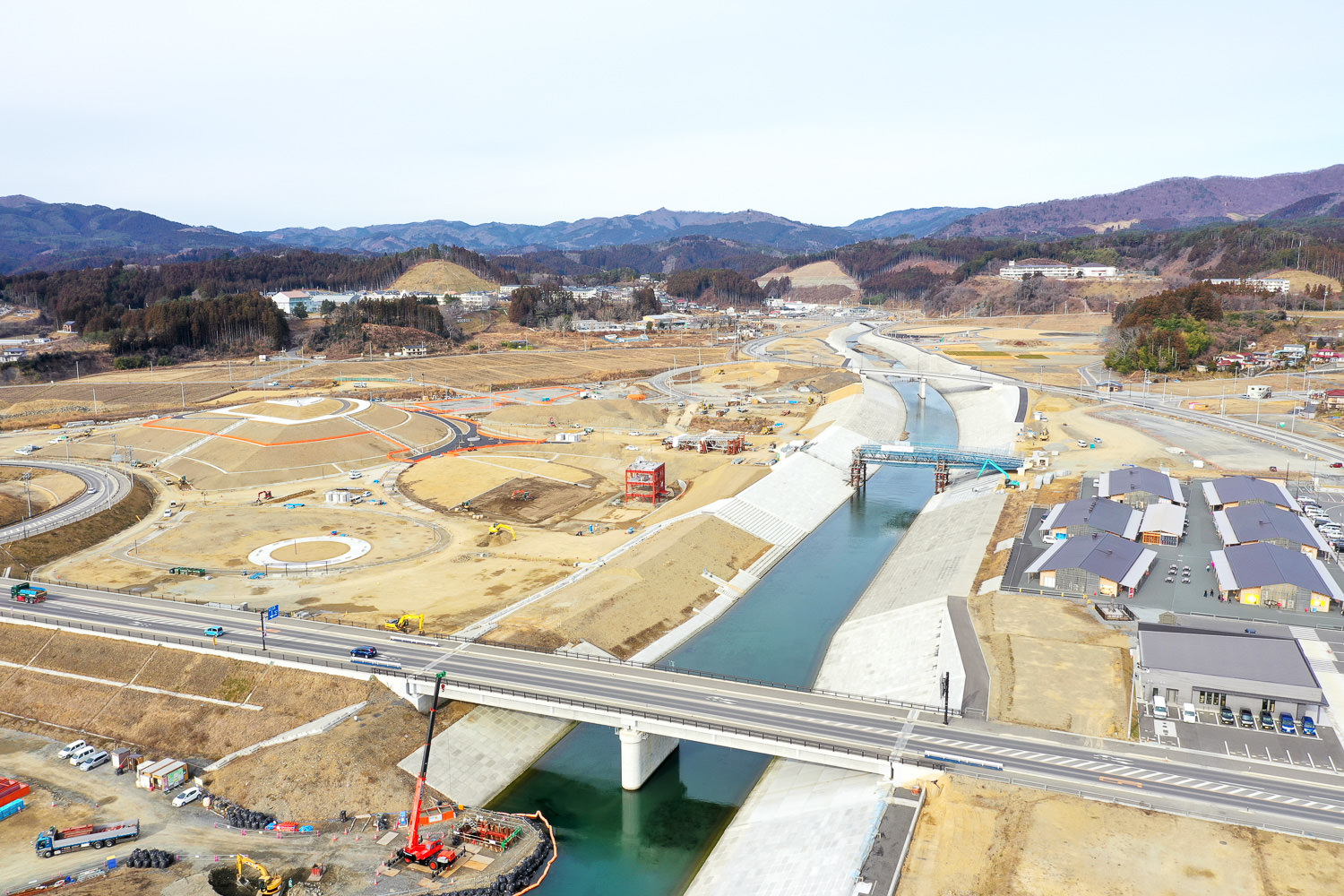 川が流れる広大な工事現場で、橋が川を横断し、複数の建設車両や機材が点在しており、周囲には道路や建物、円形の構造物が見られ、背景には丘や山々が広がっている写真