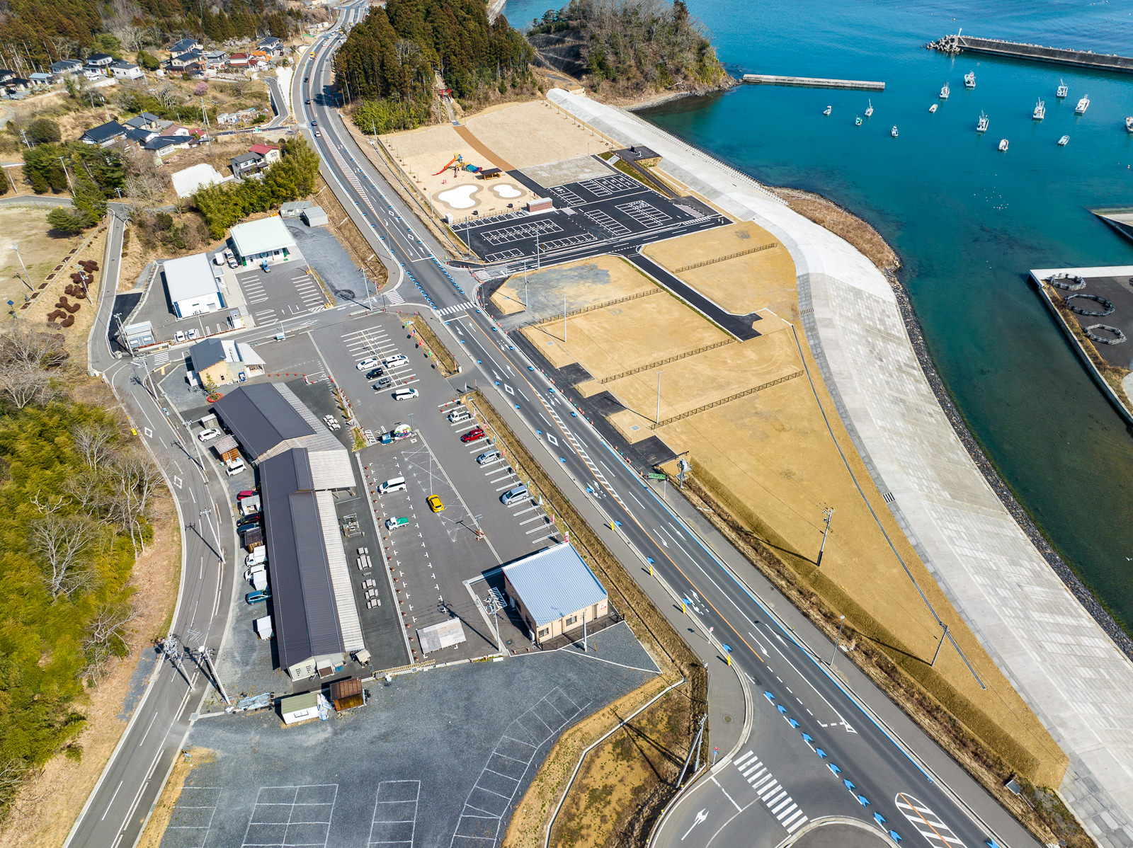 海沿いの護岸に沿って整備された道路と建物が並んだ敷地内の駐車場、公園が並び、左側には山あいの集落や森が広がり、右手には青く澄んだ海に小型漁船が点在している様子が見える、震災後の再整備が進んだ海辺の昼間の風景を高台から定点カメラで撮影した写真