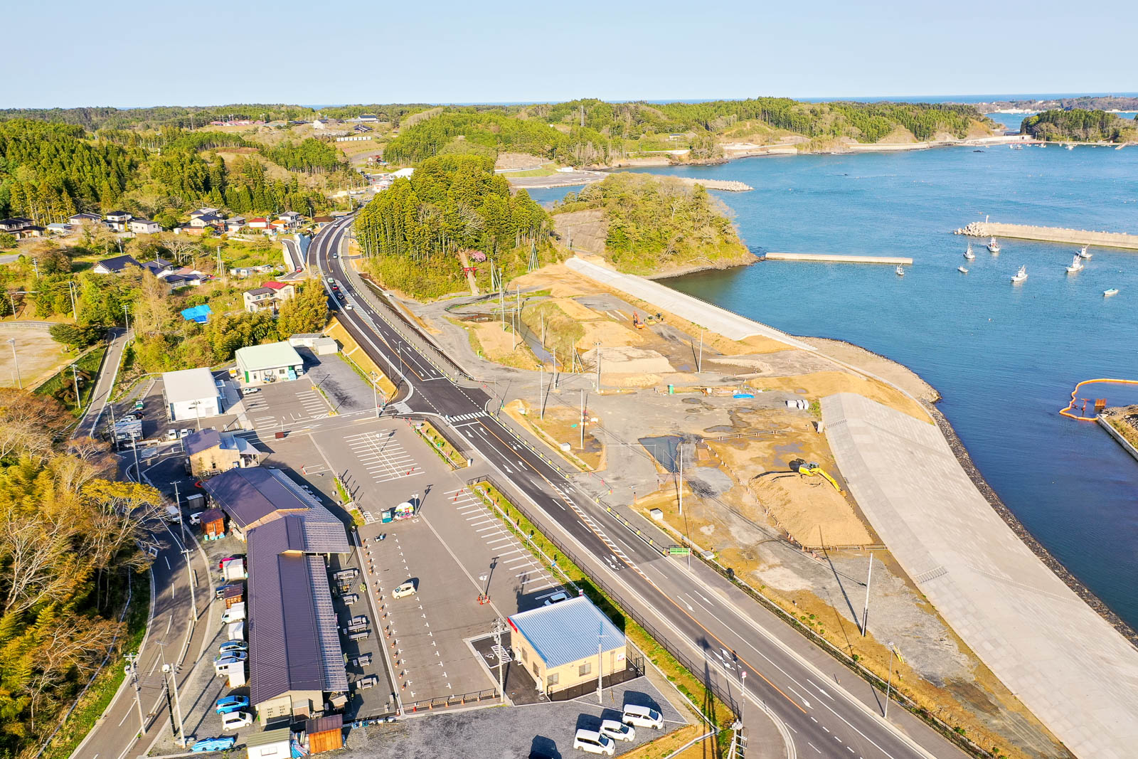 手前に広い駐車場と数棟の建物が並び、その先に新しく整備された道路がカーブを描いて海沿いを走り、右側では護岸工事が進行し、奥には穏やかな海と防波堤内に停泊する漁船、さらに小島や緑に覆われた丘が広がる様子を快晴の昼間に撮影した沿岸地域の復興中の町の写真