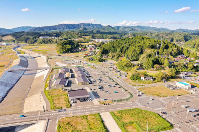 山あいの町の全景を高所から撮影していて、画面左には新しく整備された堤防と河川が見え、画面中央には複数の建物と広い駐車場があり、その奥には住宅や森林が広がっている様子がわかる写真