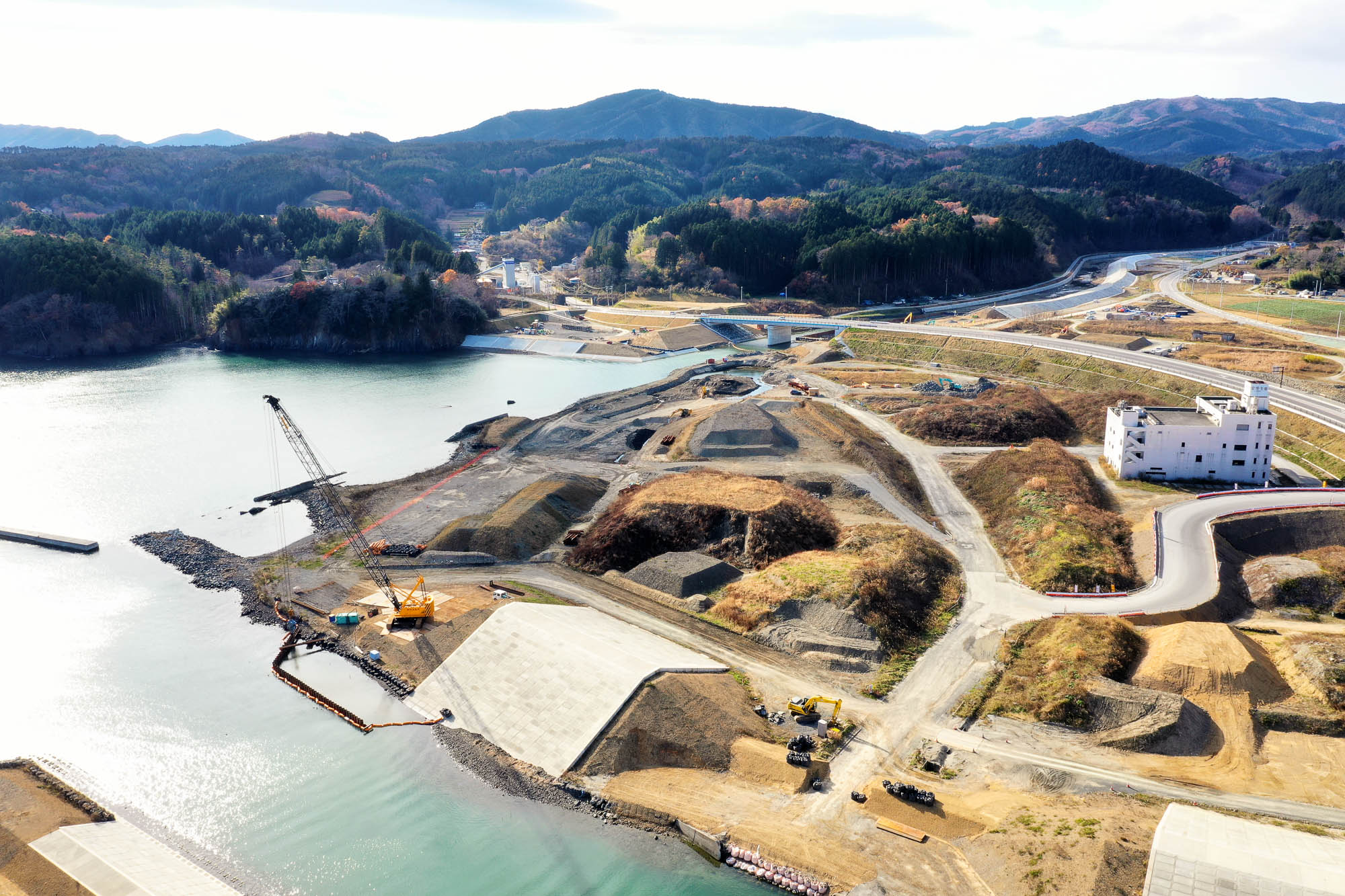 クレーン車が設置された海沿いの工事現場と、背後に山と林が広がり、曲がりくねった道路が奥へと続く様子が見える海辺の工事と山あいの町の写真