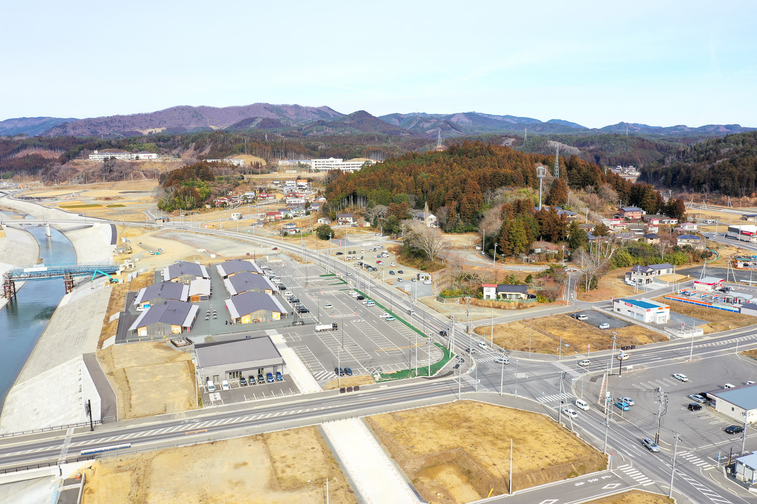 上空から撮影された小さな町または村の風景で、住宅や商業施設などの建物が点在し、道路と川が町の中を通り、背景には丘や山々が広がっている写真