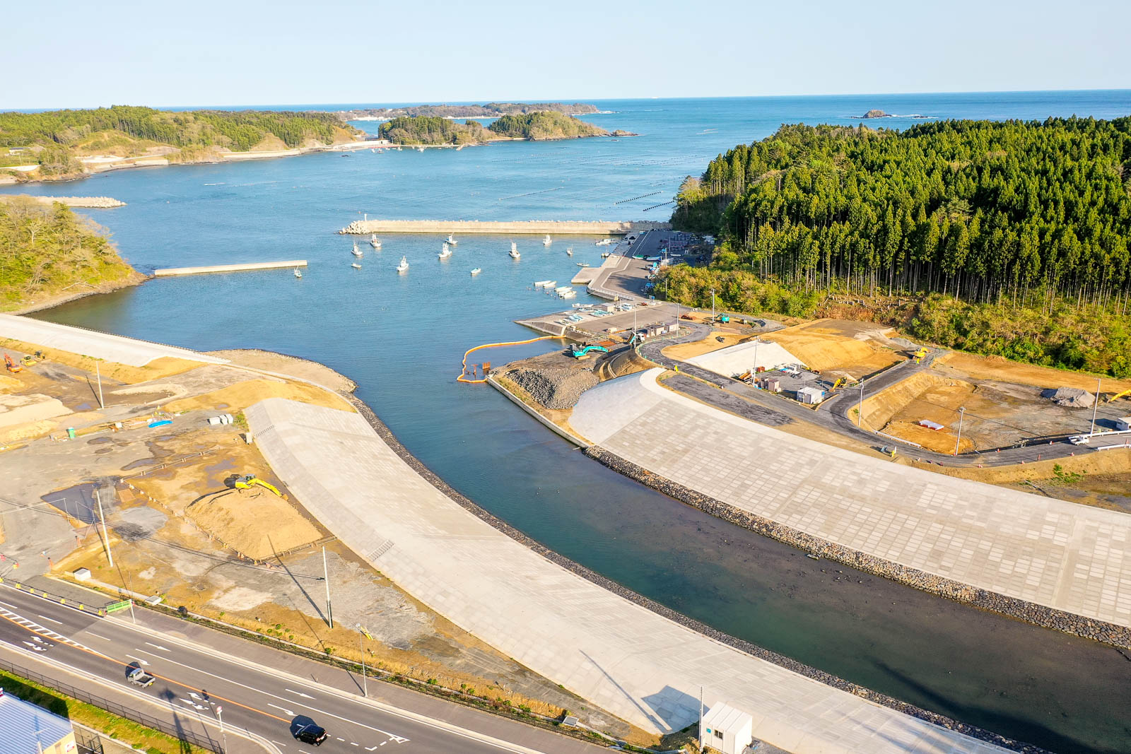 手前にコンクリートでしっかりと整備された護岸と流れる川、中央に重機が並ぶ造成地と工事現場、奥には防波堤に囲まれた港に漁船が停泊し、さらにその向こうには緑豊かな森林と小さな島々、青く広がる海が見える様子を快晴の下で撮影した沿岸部の復興工事中の港の写真