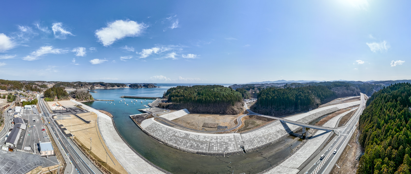 海岸線から山に囲まれた地域まで広く見渡せる、防潮堤と川の護岸整備が進む湾に面した小さな町の昼間の風景を高台から定点カメラで撮影した写真