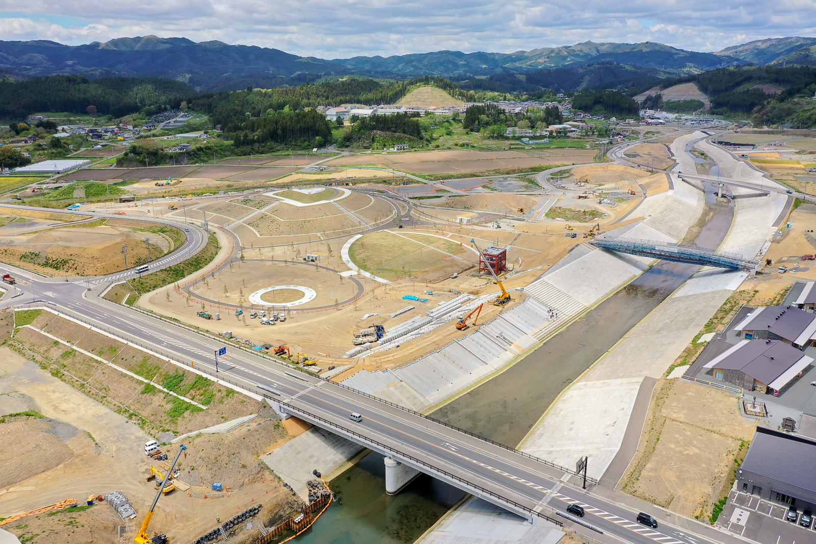 川または運河に架かる橋を中心に、周囲で円形や曲線状の構造物が建設されており、建設機材や車両が点在し、背景には山々と緑が広がる開発中の地域を空から見下ろした橋と周辺工事の写真