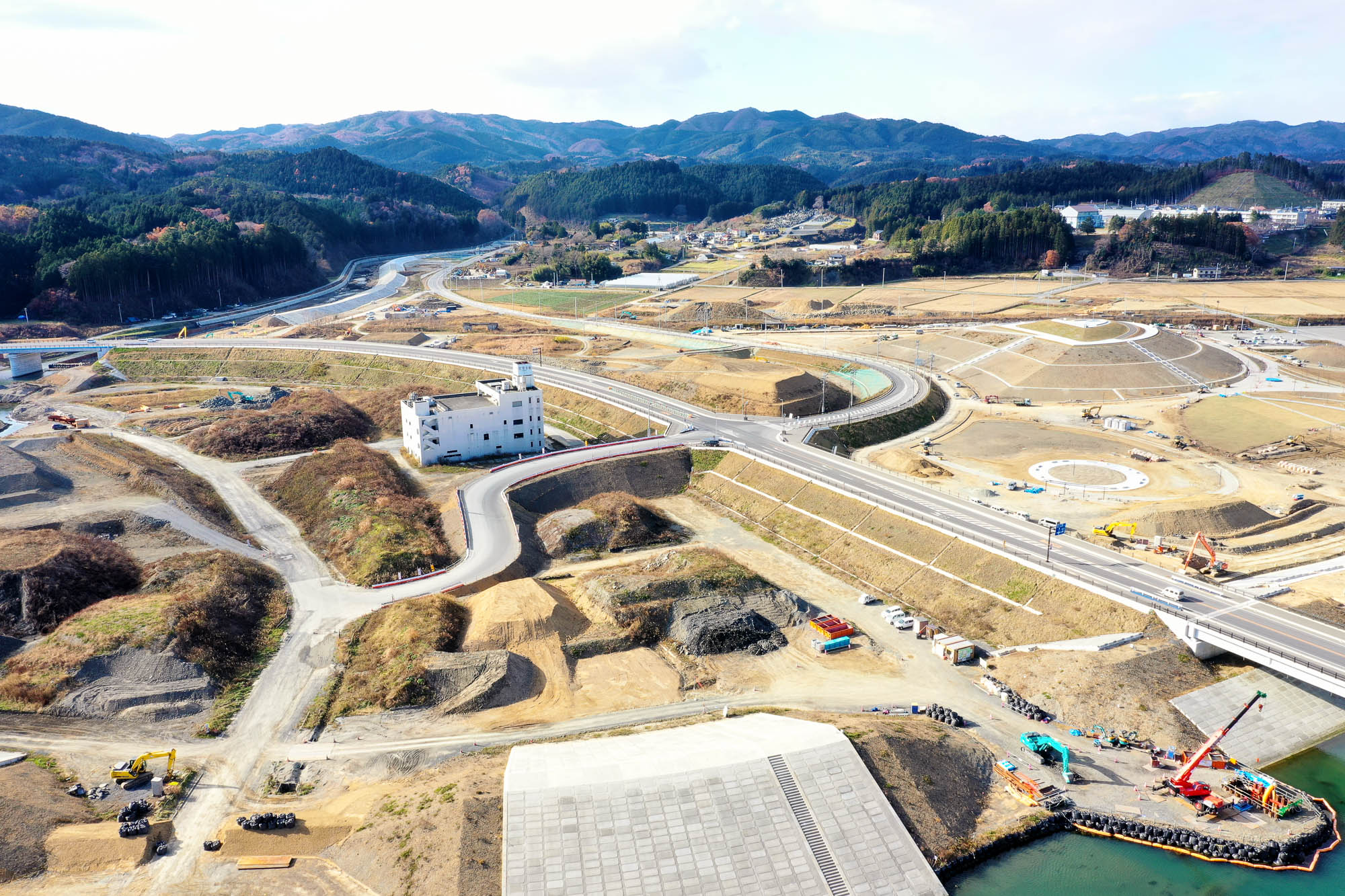白い建物と高架道路を中心に、複雑に交差する道路や造成地が広がり、奥には山並みに囲まれた町が広がっている高架道路と造成地の写真